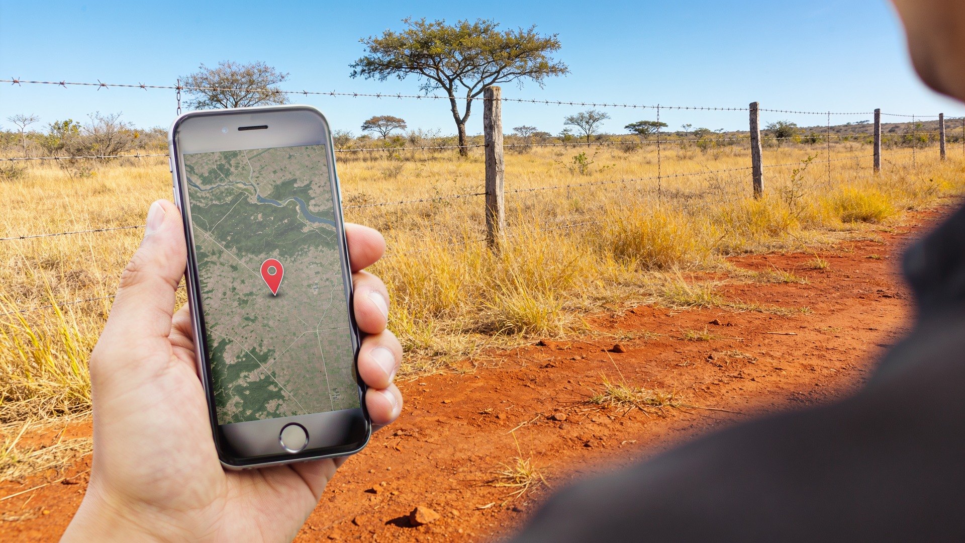 Mão segurando um celular com mapa e pino de localização em um pasto seco — o GPS mostra onde você está, não a água que existe embaixo (imagem ilustrativa)