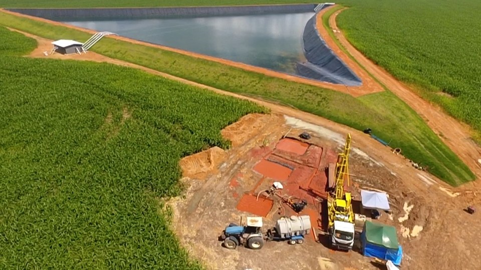 Vista aérea de um canteiro de perfuração de poço tubular, com a sonda e os equipamentos em solo vermelho, ao lado de um reservatório revestido com geomembrana no meio de uma grande lavoura verde